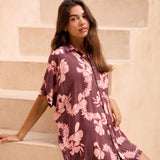 woman with brown hair wearing.a brown/maroon button up dress with pink lei awapuhi print