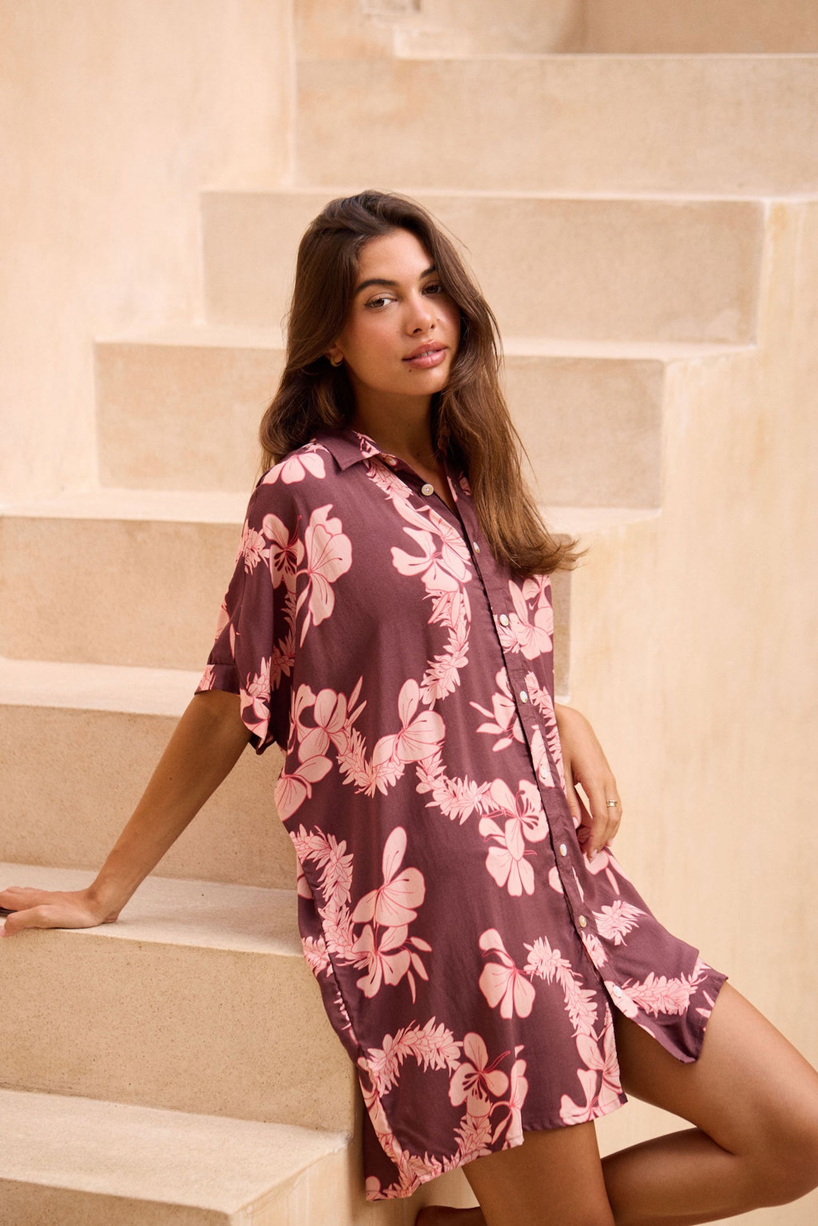woman with brown hair wearing.a brown/maroon button up dress with pink lei awapuhi print