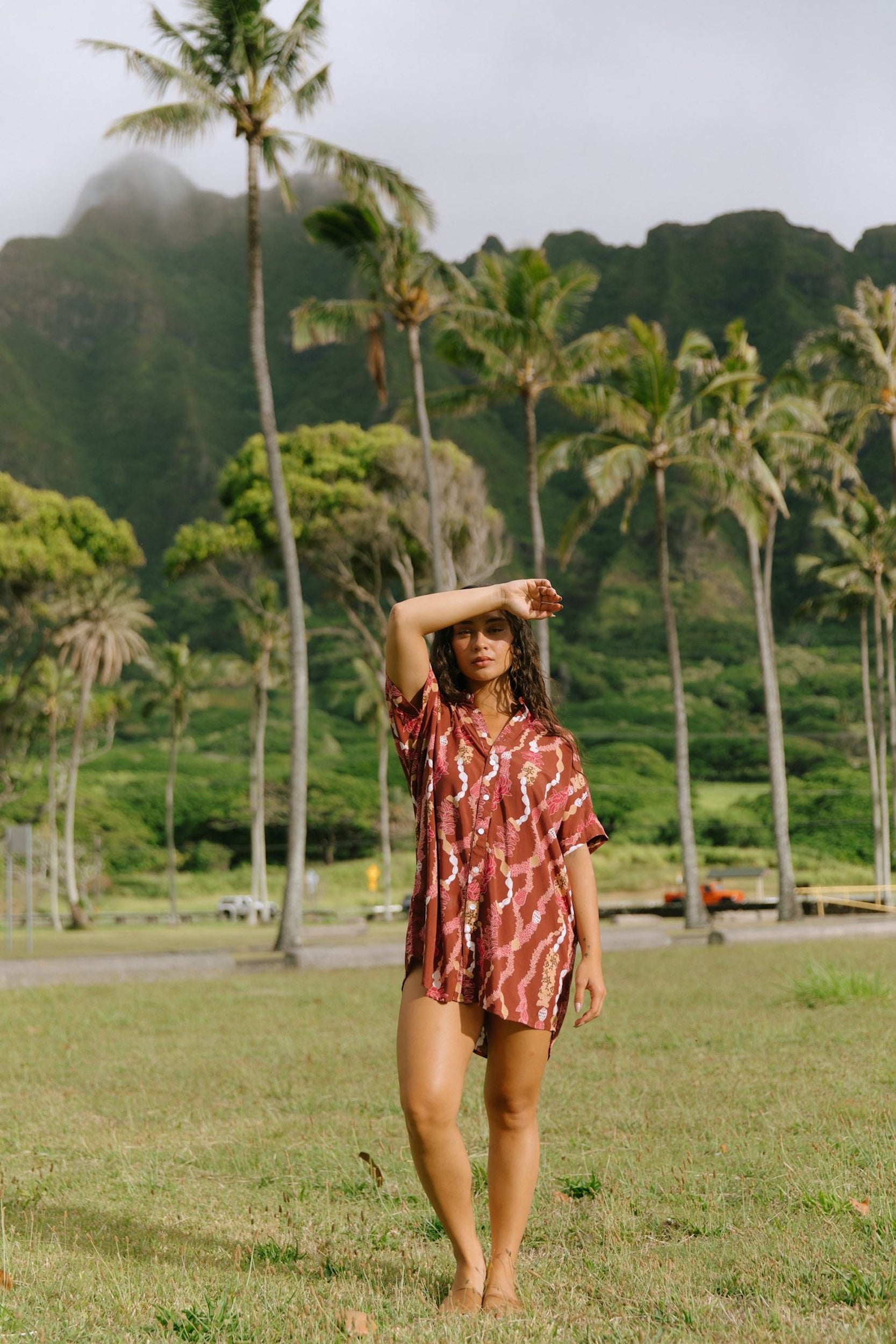 woman with brown hair wearing a red-brown colored button up t-shirt dress with lei print
