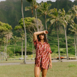 woman with brown hair wearing a red-brown colored button up t-shirt dress with lei print
