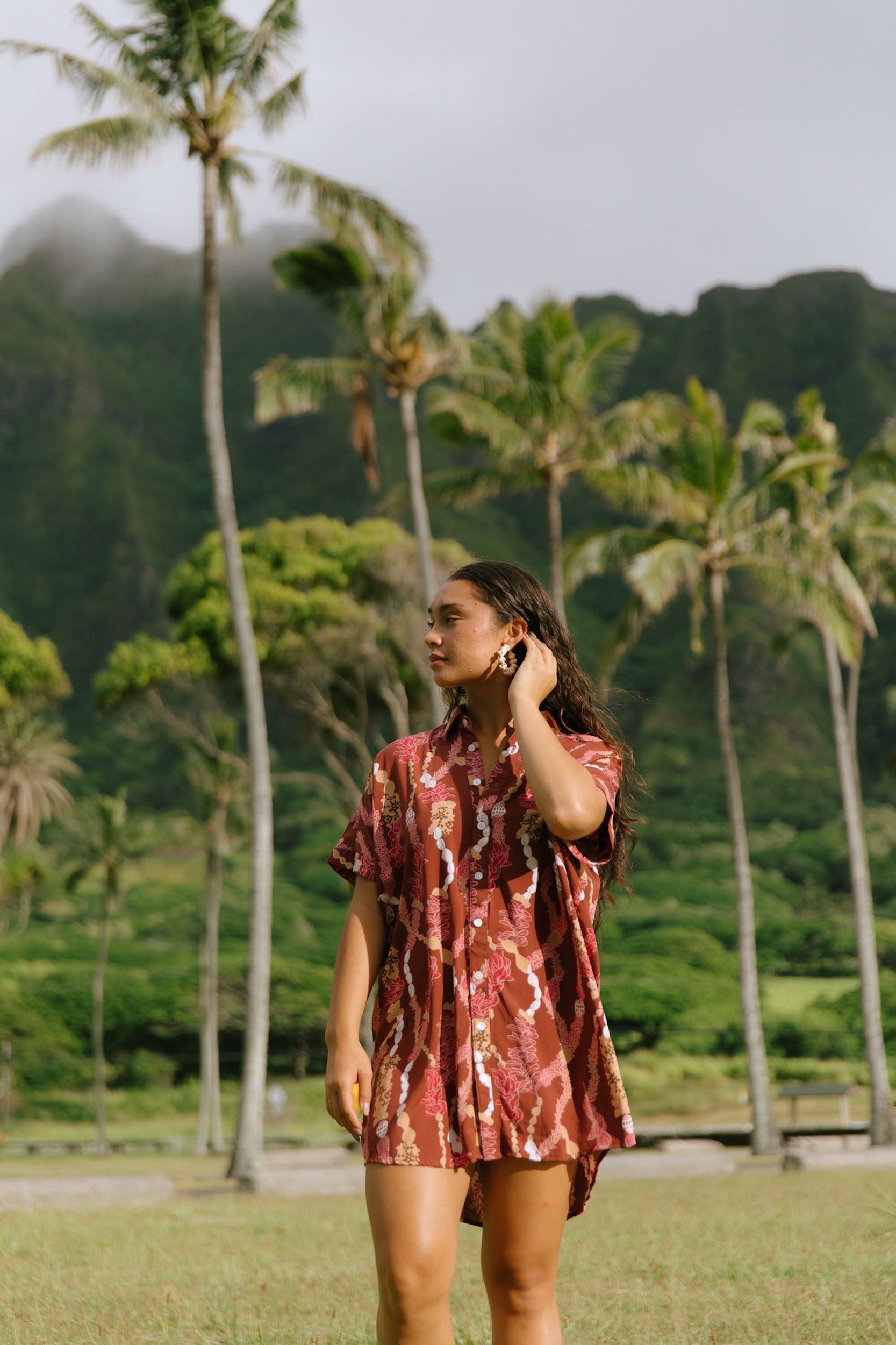 woman with brown hair wearing a red-brown colored button up t-shirt dress with lei print