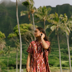 woman with brown hair wearing a red-brown colored button up t-shirt dress with lei print