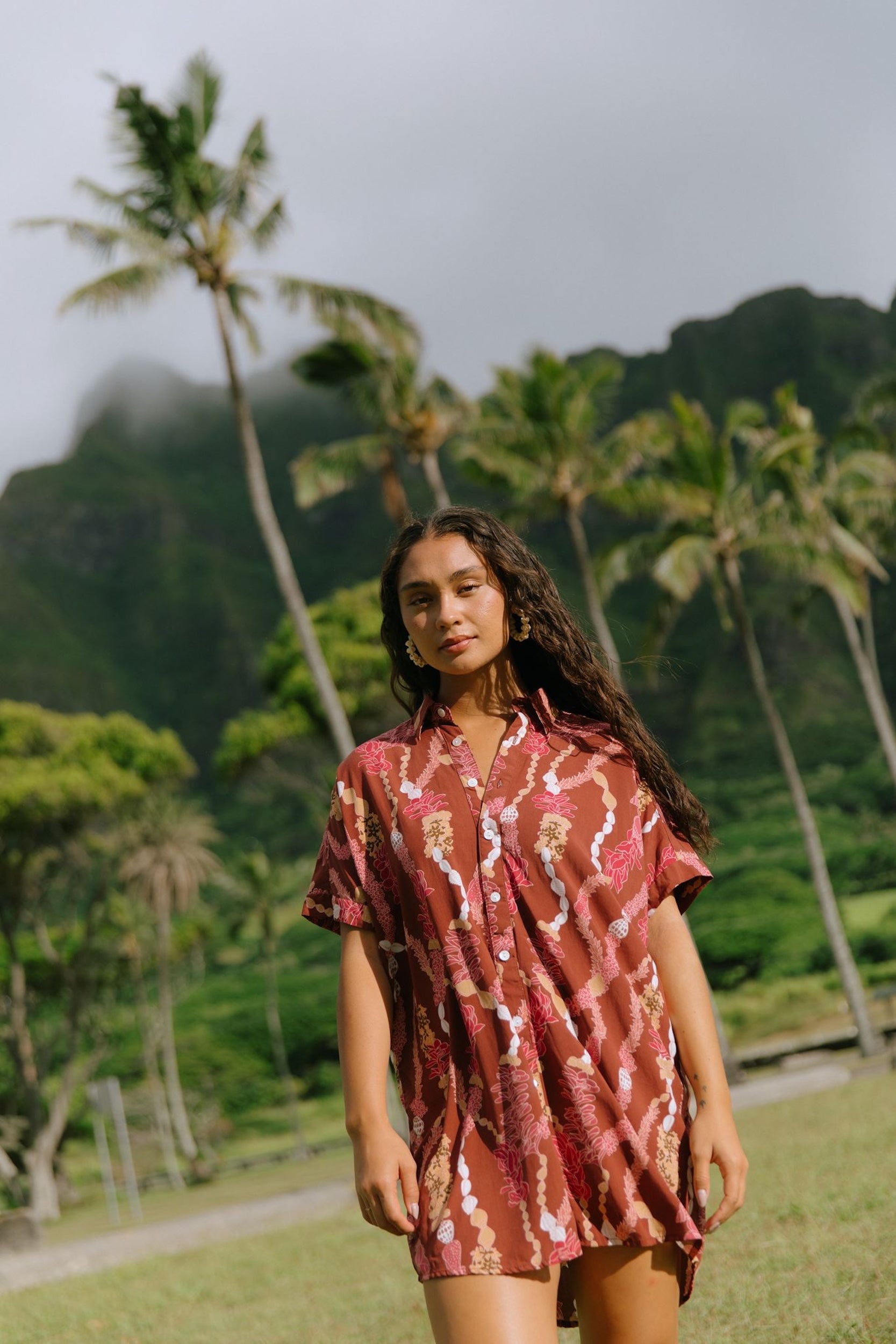 woman with brown hair wearing a red-brown colored button up t-shirt dress with lei print