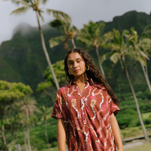 woman with brown hair wearing a red-brown colored button up t-shirt dress with lei print