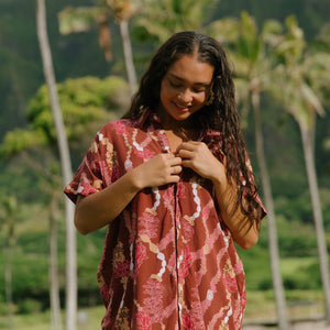 woman with brown hair wearing a red-brown colored button up t-shirt dress with lei print