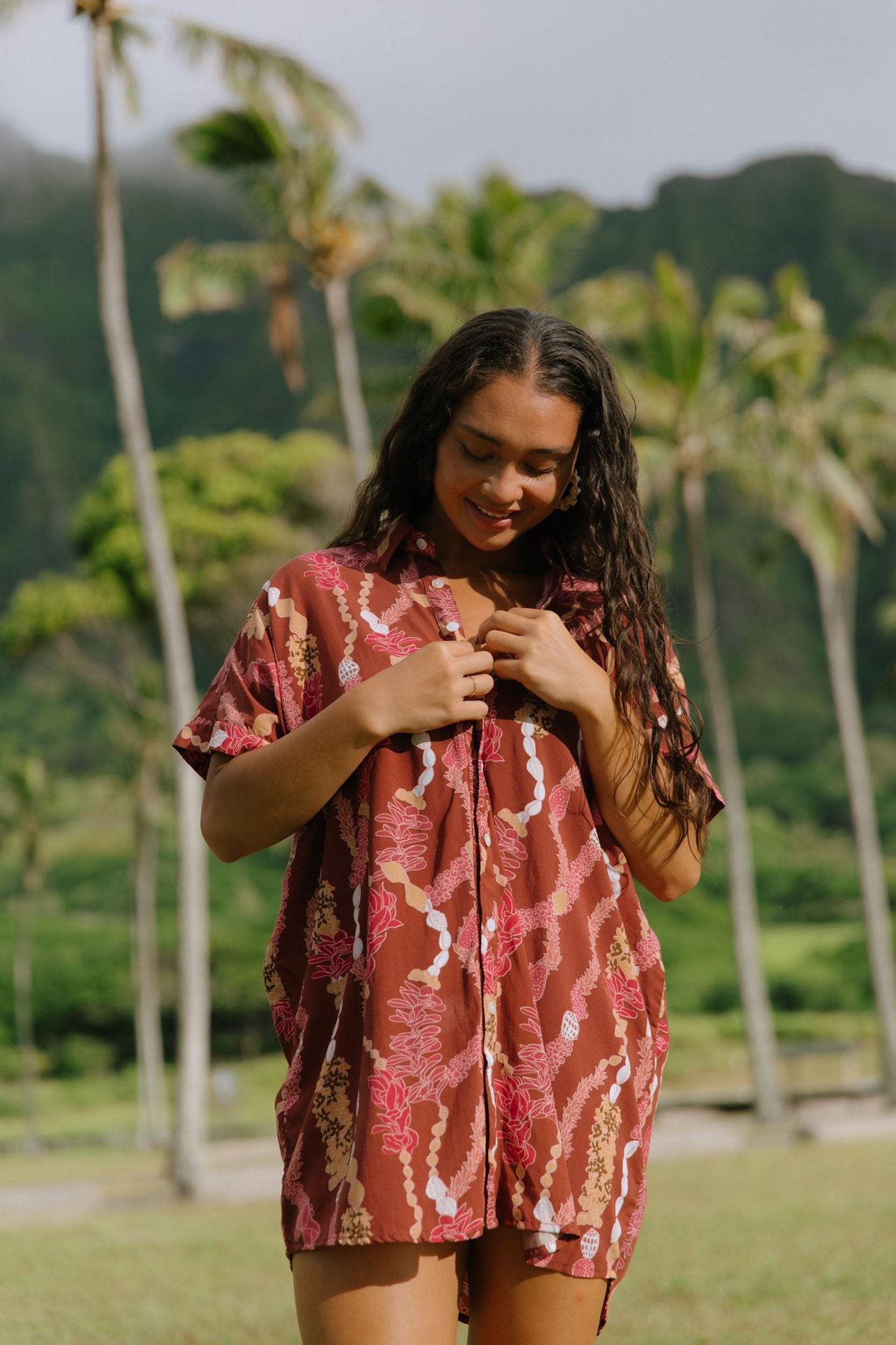 woman with brown hair wearing a red-brown colored button up t-shirt dress with lei print