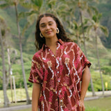 woman with brown hair wearing a red-brown colored button up t-shirt dress with lei print