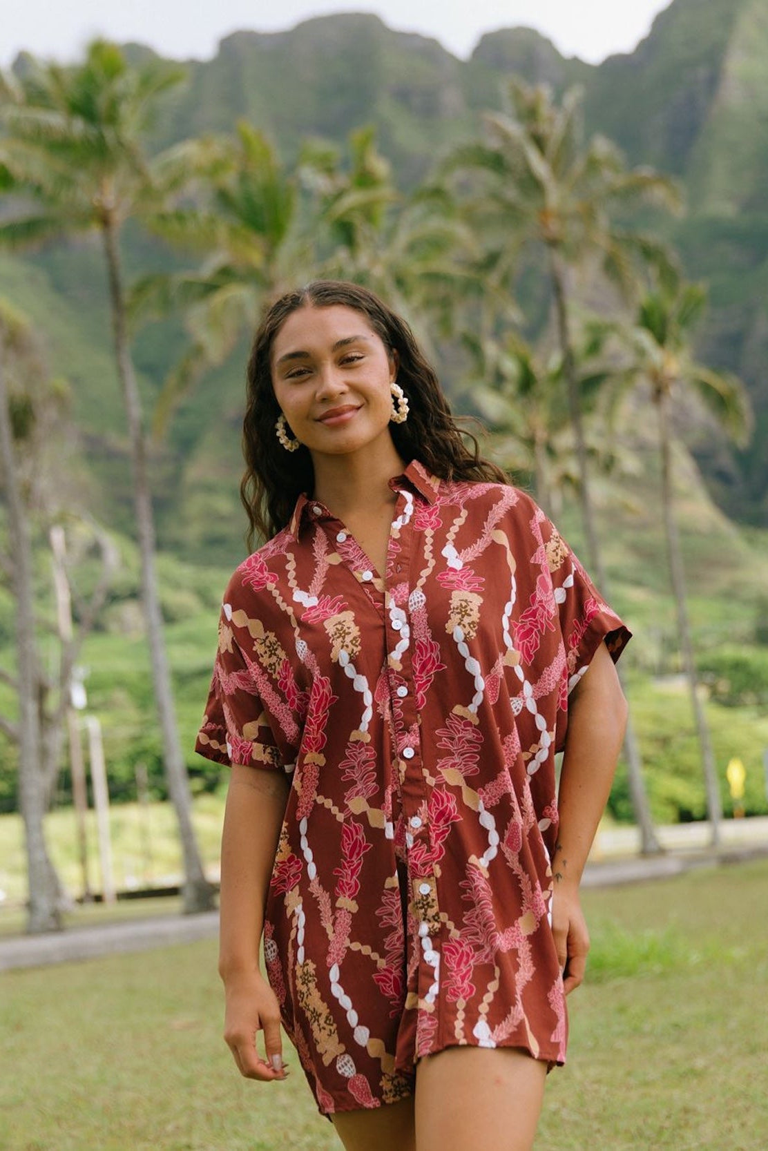 woman with brown hair wearing a red-brown colored button up t-shirt dress with lei print
