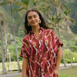woman with brown hair wearing a red-brown colored button up t-shirt dress with lei print
