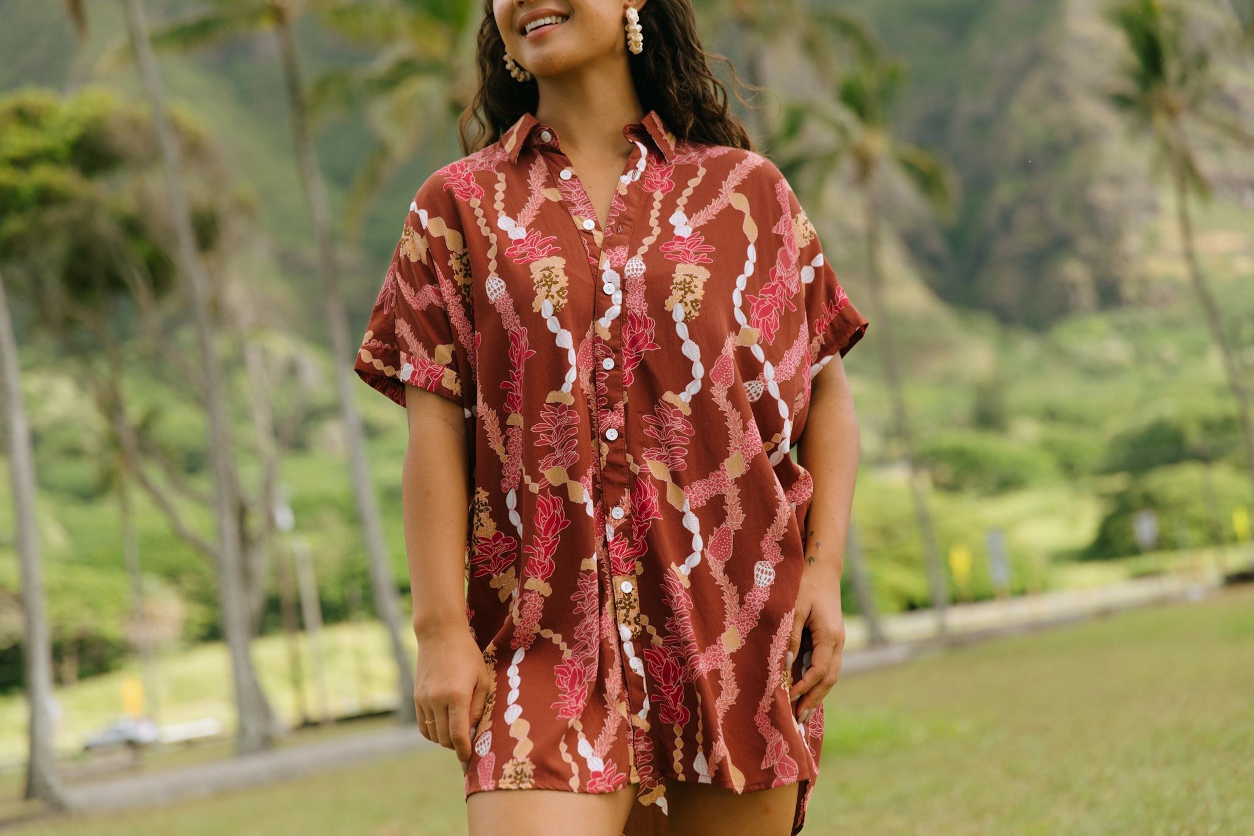 woman with brown hair wearing a red-brown colored button up t-shirt dress with lei print