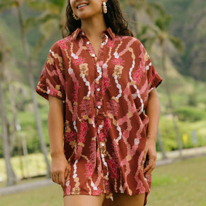 woman with brown hair wearing a red-brown colored button up t-shirt dress with lei print