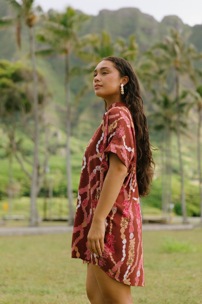 woman with brown hair wearing a red-brown colored button up t-shirt dress with lei print