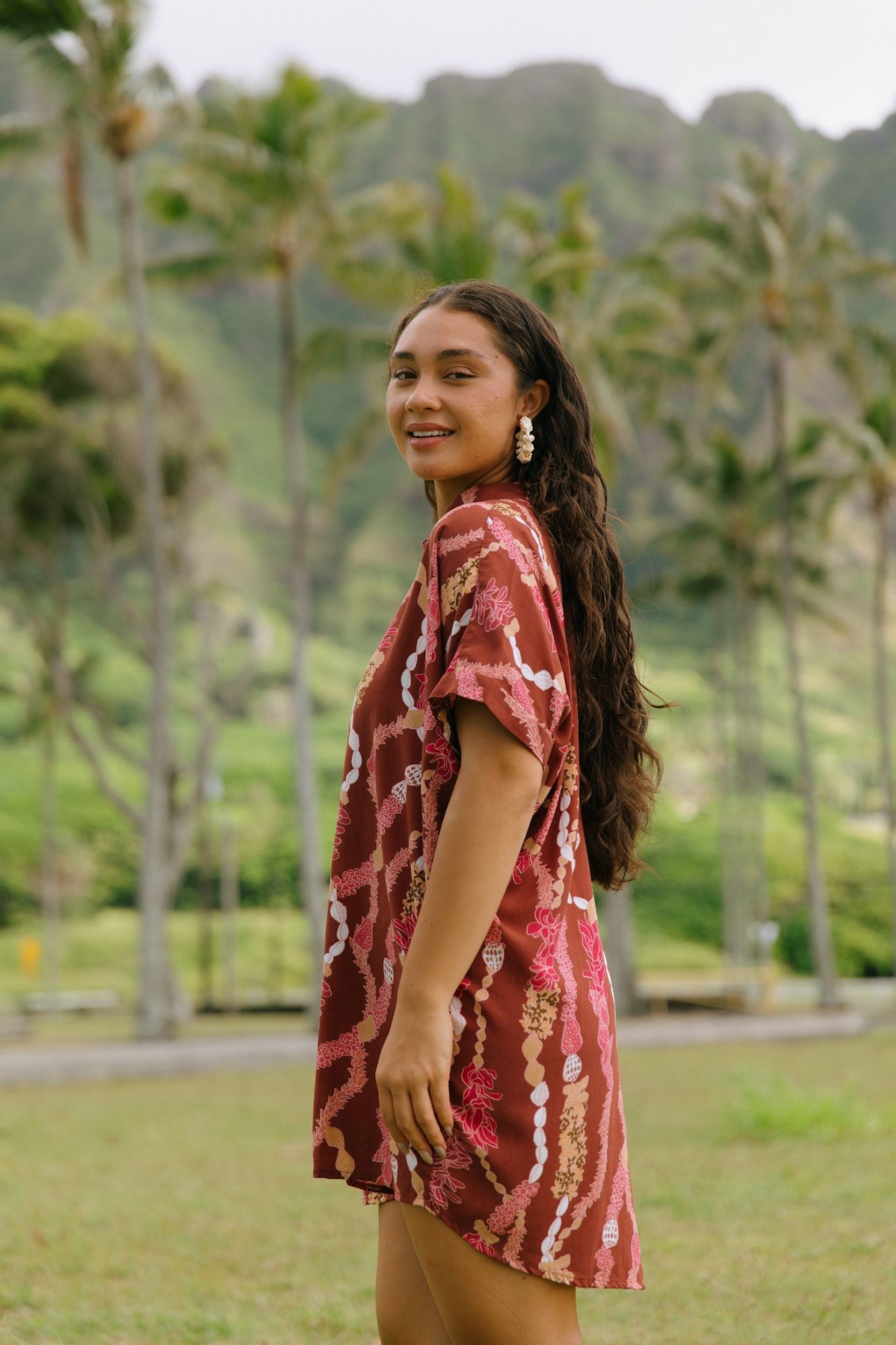 woman with brown hair wearing a red-brown colored button up t-shirt dress with lei print