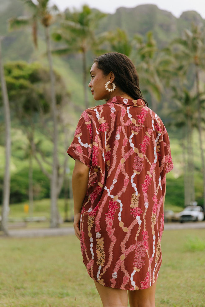woman with brown hair wearing a red-brown colored button up t-shirt dress with lei print