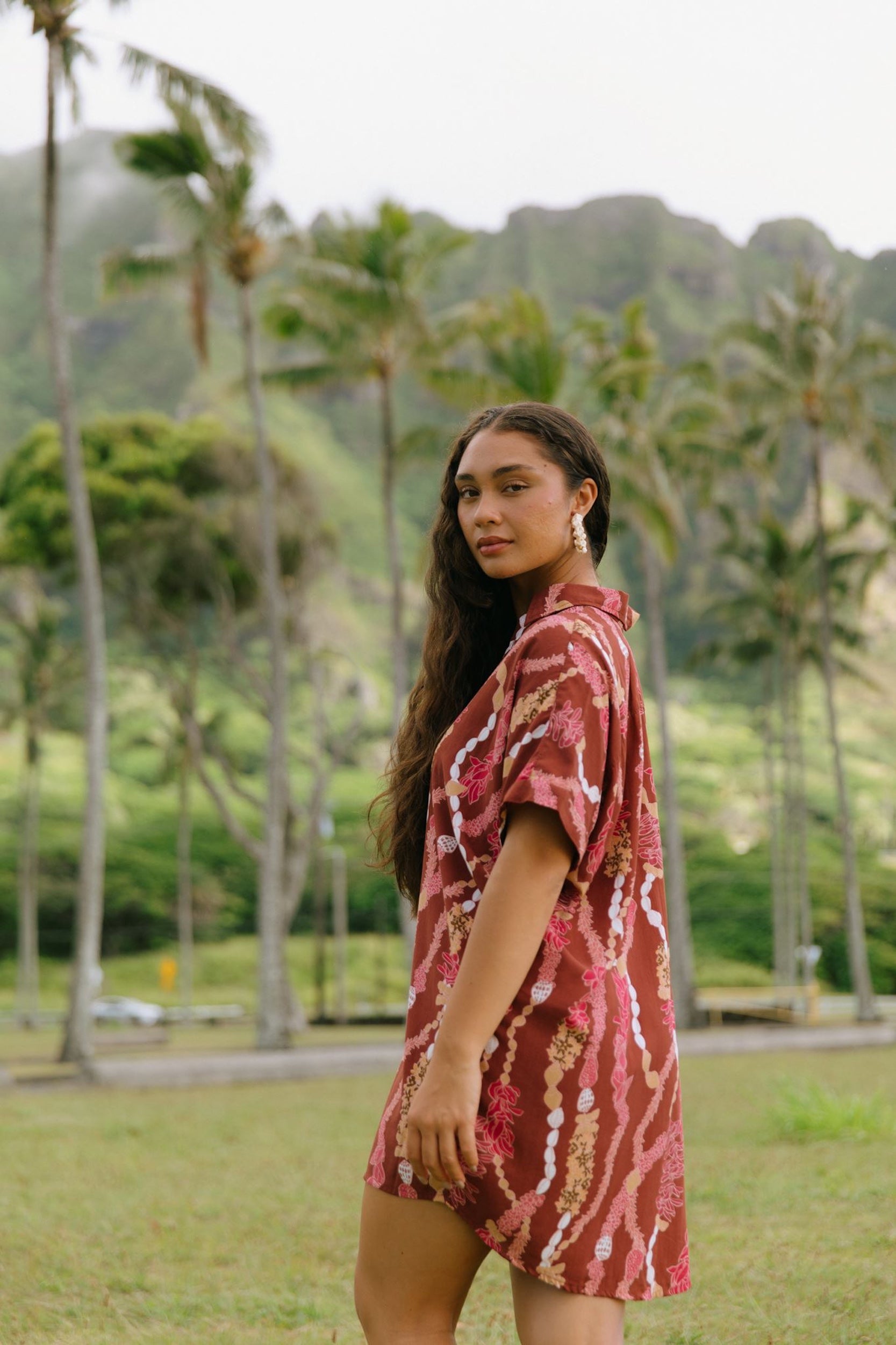 woman with brown hair wearing a red-brown colored button up t-shirt dress with lei print