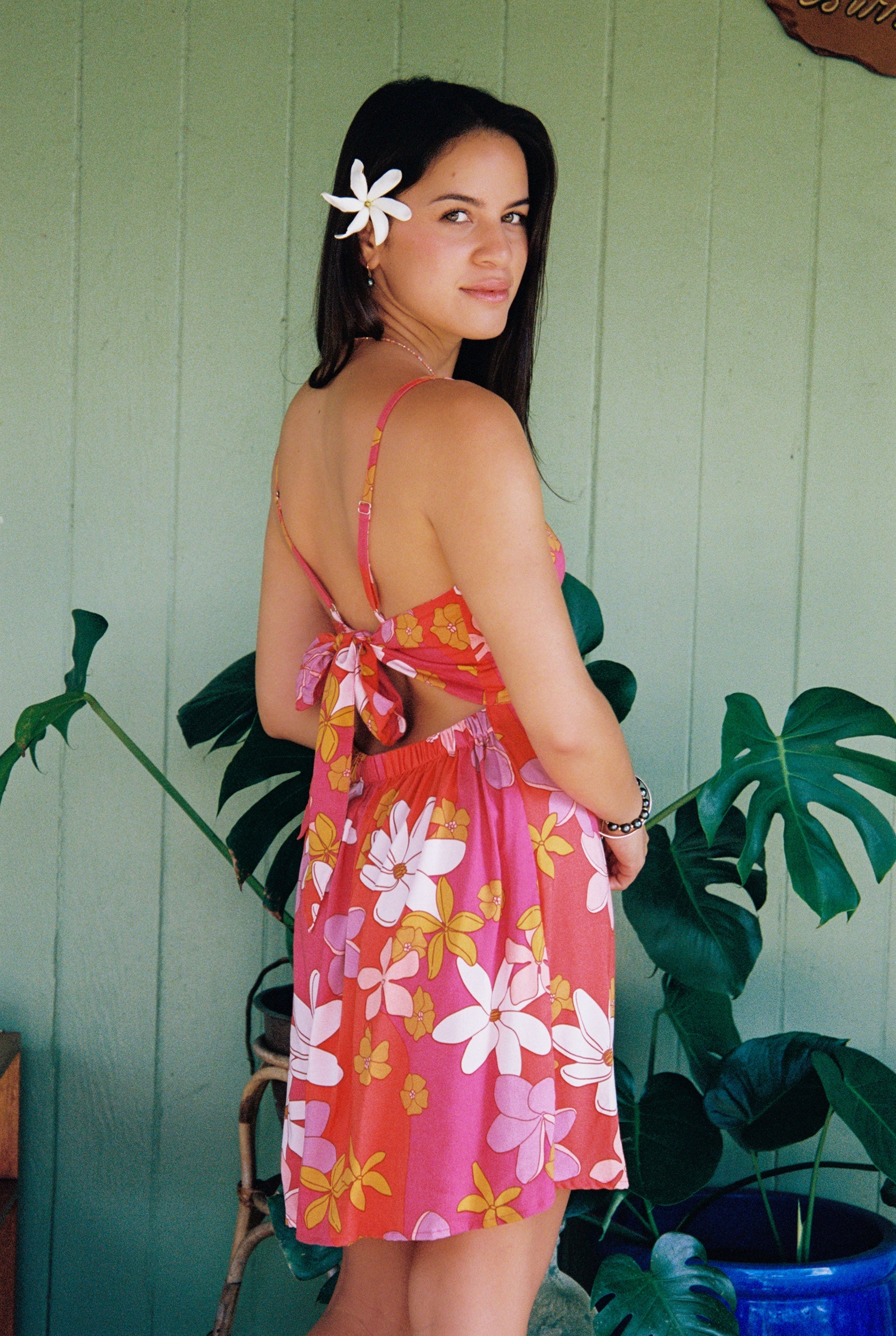 woman with brown hair wearing a pink mini spaghetti strap dress with retro flower print and tie back detail 