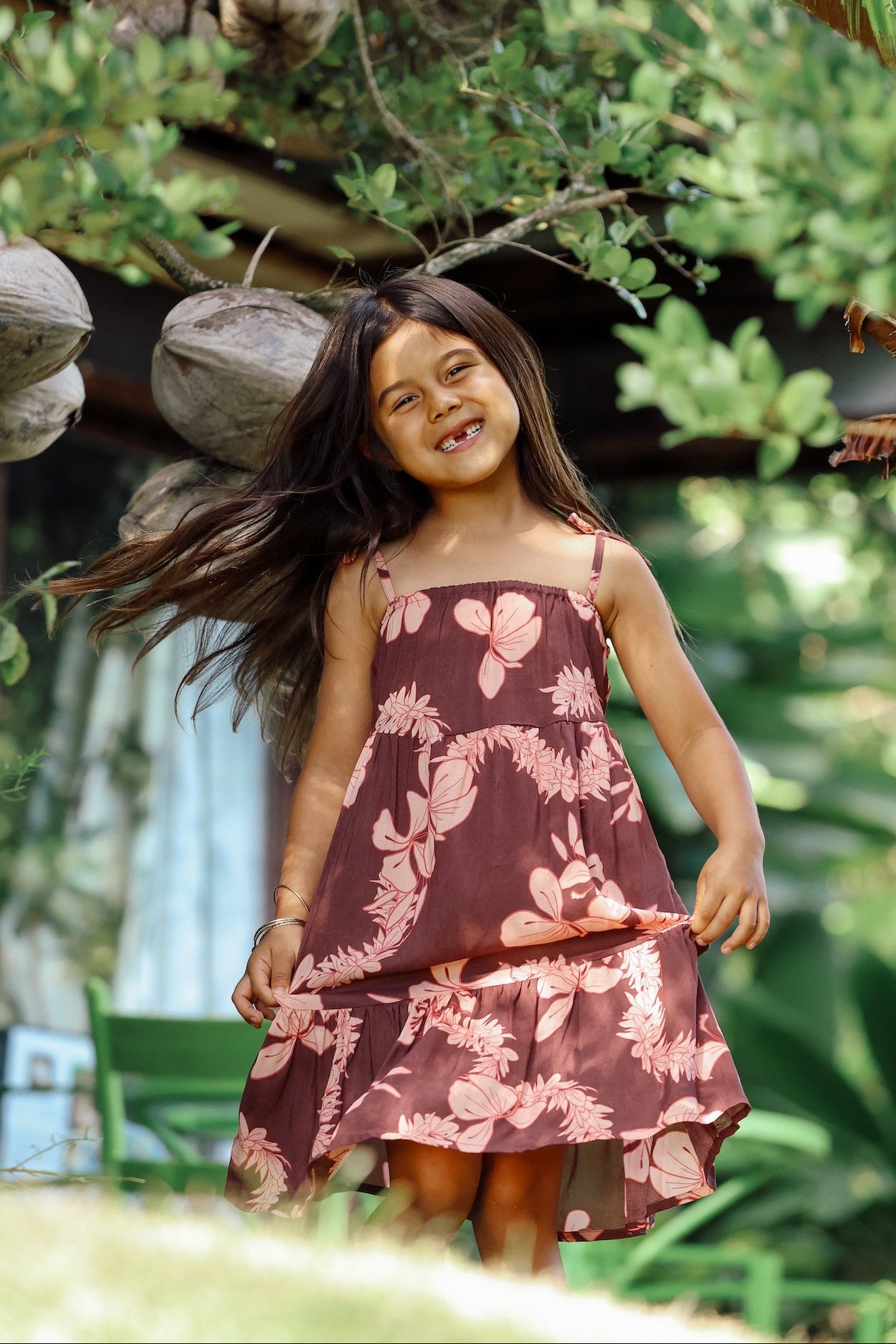 little girl wearing a long brown dress with tie sleeve with light pink lei awapuhi print