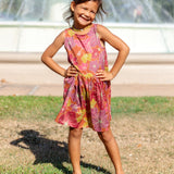 little girl wearing terracotta colored high neck sleeveless knee length dress with large purple and yellow flower print