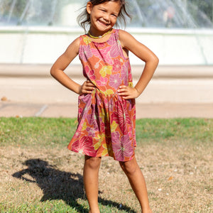 little girl wearing terracotta colored high neck sleeveless knee length dress with large purple and yellow flower print