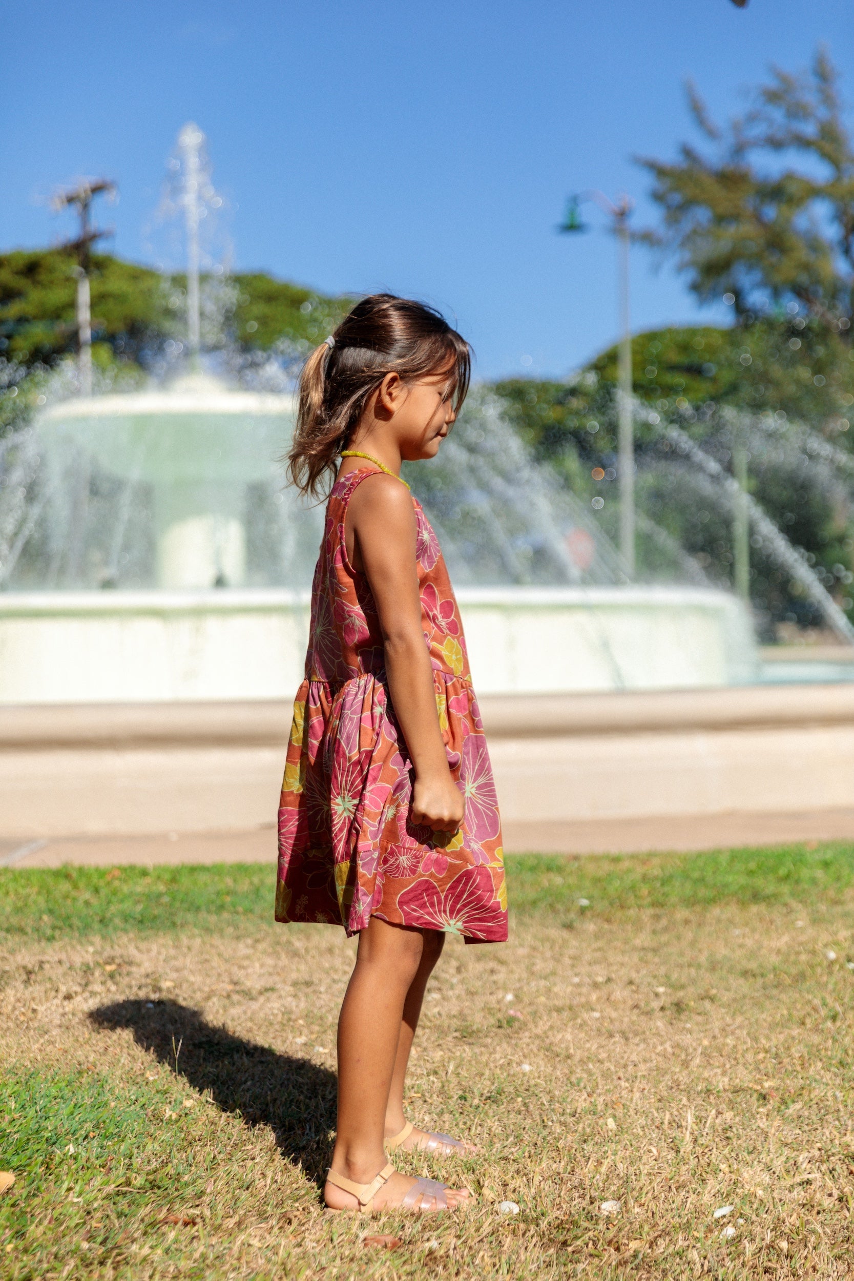 little girl wearing terracotta colored high neck sleeveless knee length dress with large purple and yellow flower print