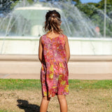 little girl wearing terracotta colored high neck sleeveless knee length dress with large purple and yellow flower print