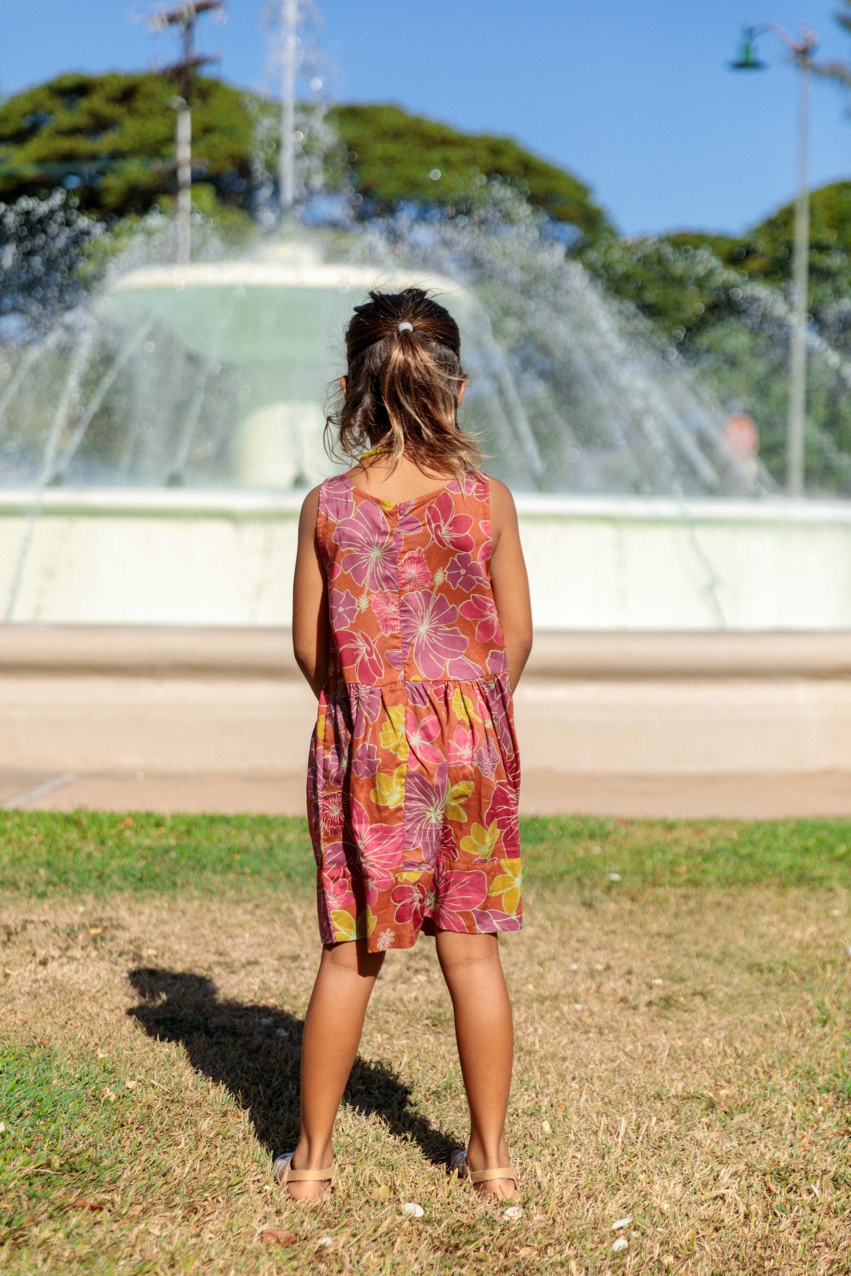 little girl wearing terracotta colored high neck sleeveless knee length dress with large purple and yellow flower print