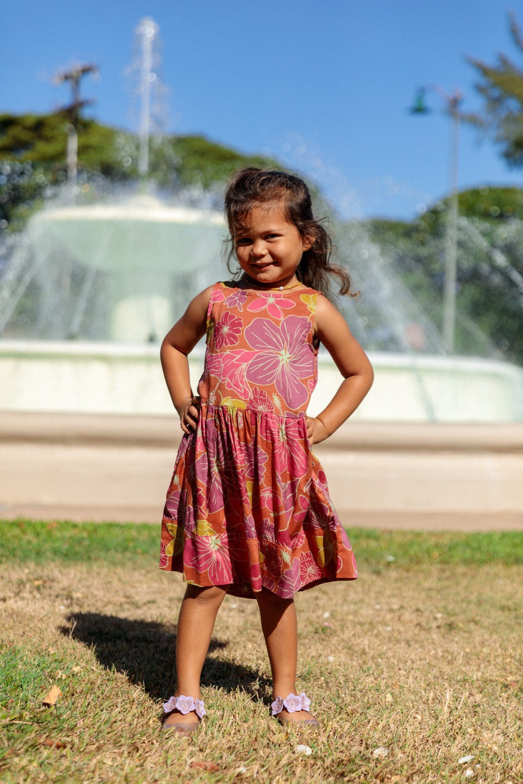 little girl wearing terracotta colored high neck sleeveless knee length dress with large purple and yellow flower print