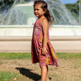 little girl wearing terracotta colored high neck sleeveless knee length dress with large purple and yellow flower print