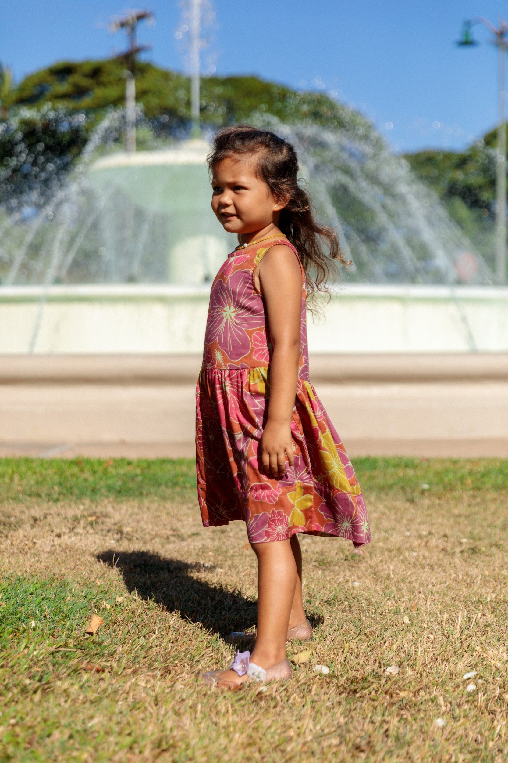little girl wearing terracotta colored high neck sleeveless knee length dress with large purple and yellow flower print