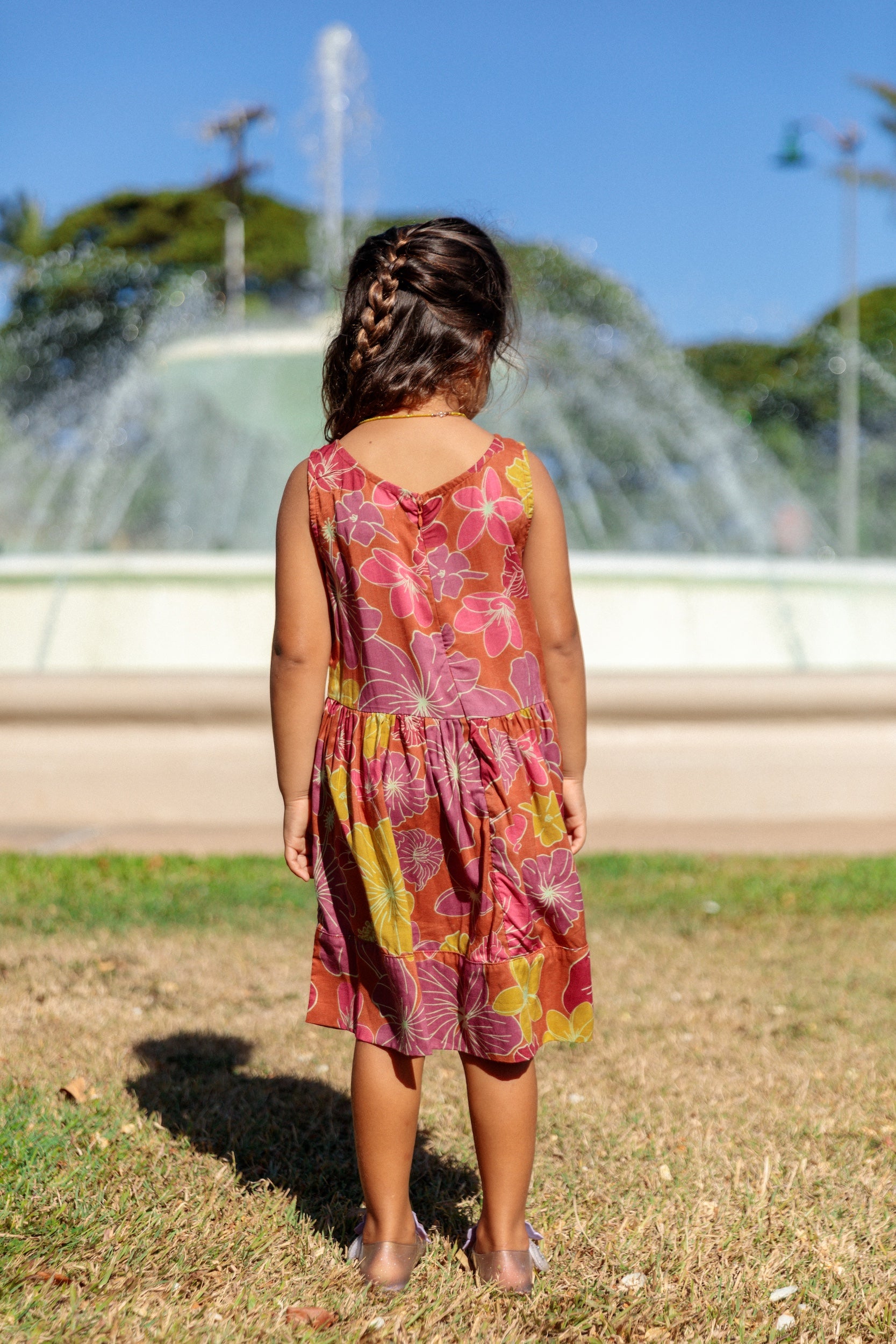 little girl wearing terracotta colored high neck sleeveless knee length dress with large purple and yellow flower print
