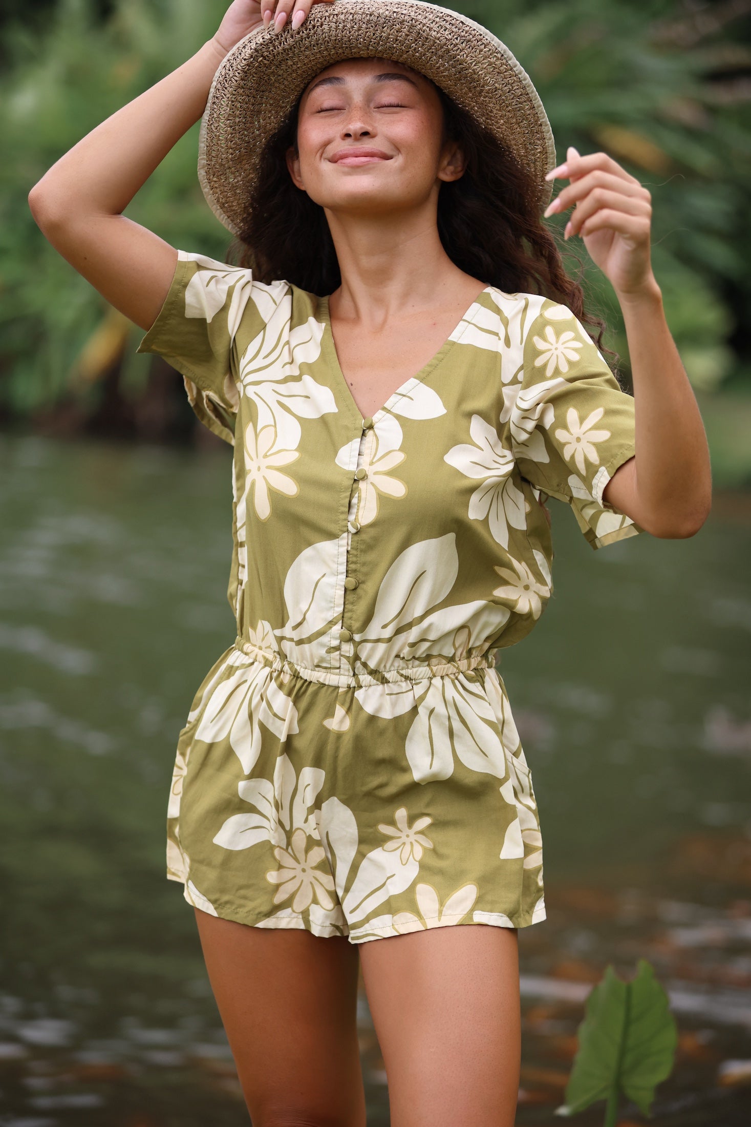 woman with brown hair wearing a short sleeve playsuit in a green color with naupaka pattern throughout. 