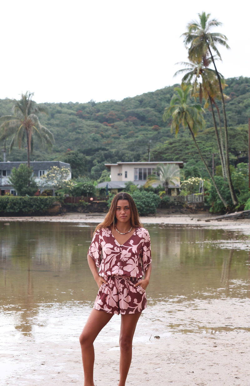 Woman with brown hair wearing a flowy button up playsuit in blush and brown hibiscus print.