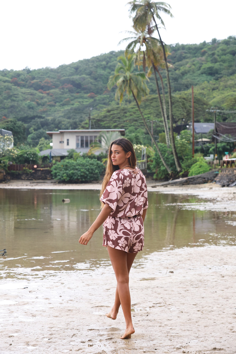 Woman with brown hair wearing a flowy button up playsuit in blush and brown hibiscus print.