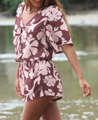 Woman with brown hair wearing a flowy button up playsuit in blush and brown hibiscus print.
