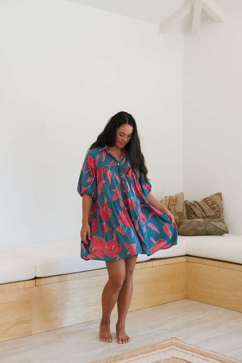 Woman wearing short loose blue dress with elbow length sleeves and large pink and red floral print.