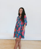 Woman wearing short loose blue dress with elbow length sleeves and large pink and red floral print.