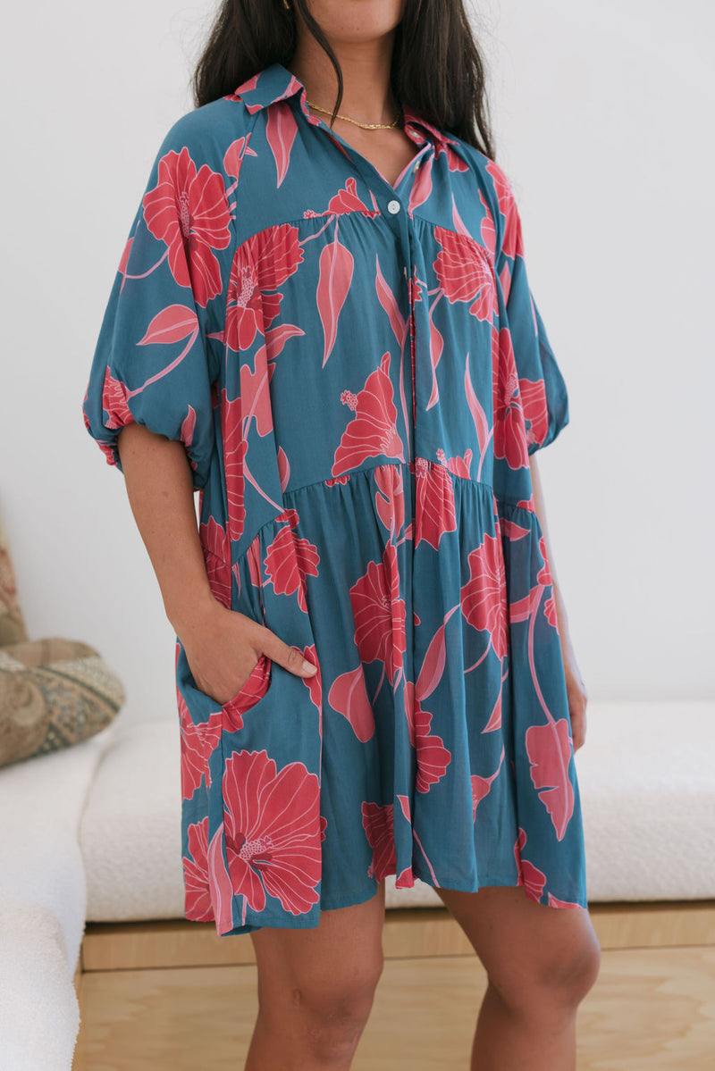 Woman wearing short loose blue dress with elbow length sleeves and large pink and red floral print.