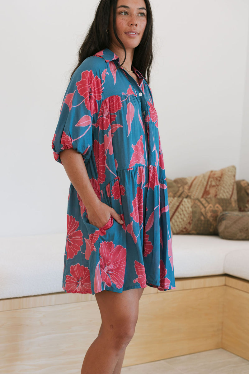 Woman wearing short loose blue dress with elbow length sleeves and large pink and red floral print.