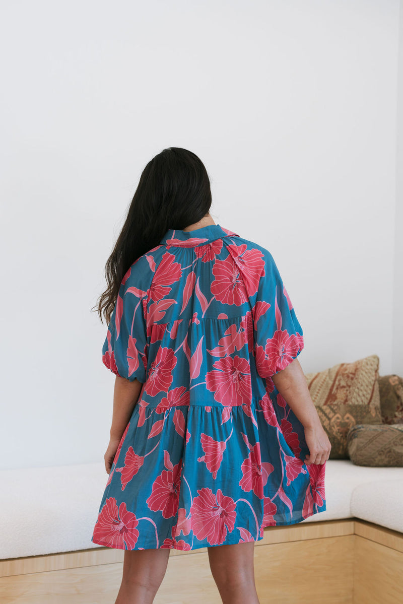 Woman wearing short loose blue dress with elbow length sleeves and large pink and red floral print.