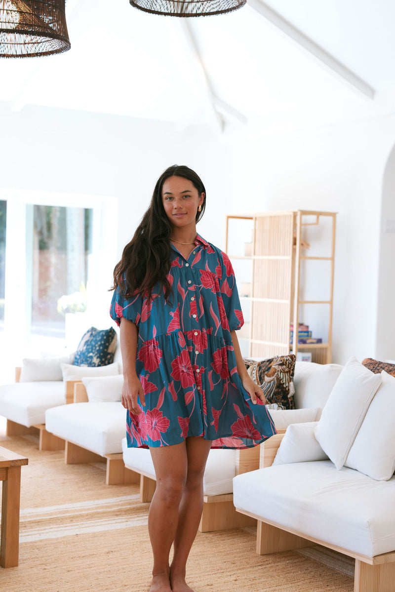 Woman wearing short loose blue dress with elbow length sleeves and large pink and red floral print.