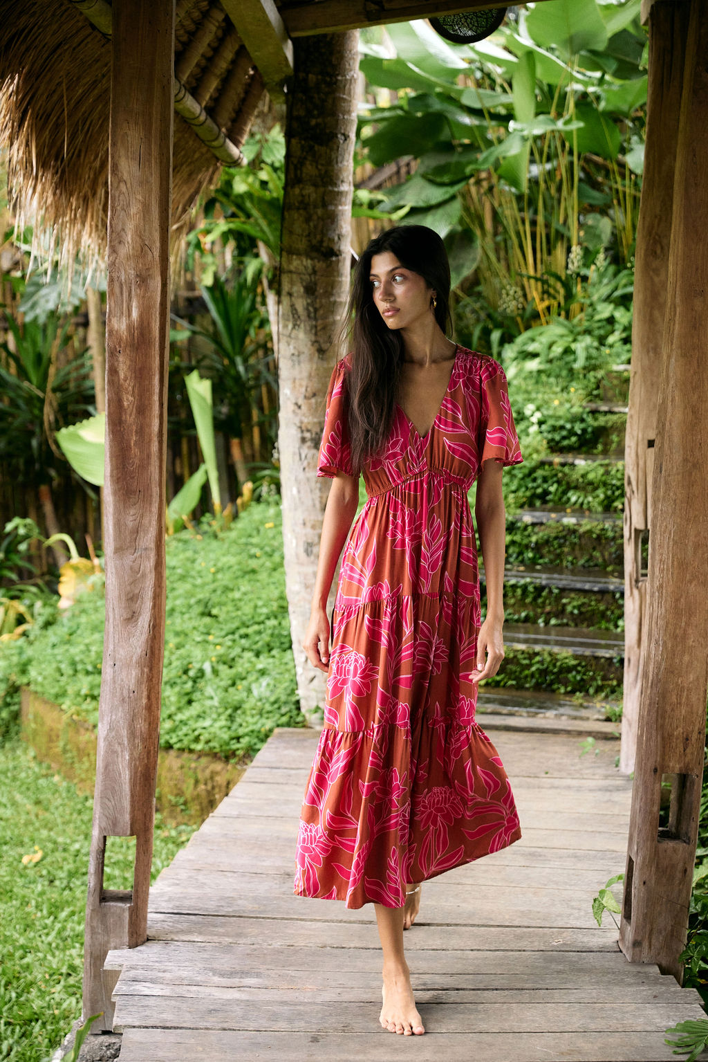 Woman with dark hair wearing tiered maxi dress with sleeves. It is the Mariana Dress in the Garden 'Awapuhi print. 