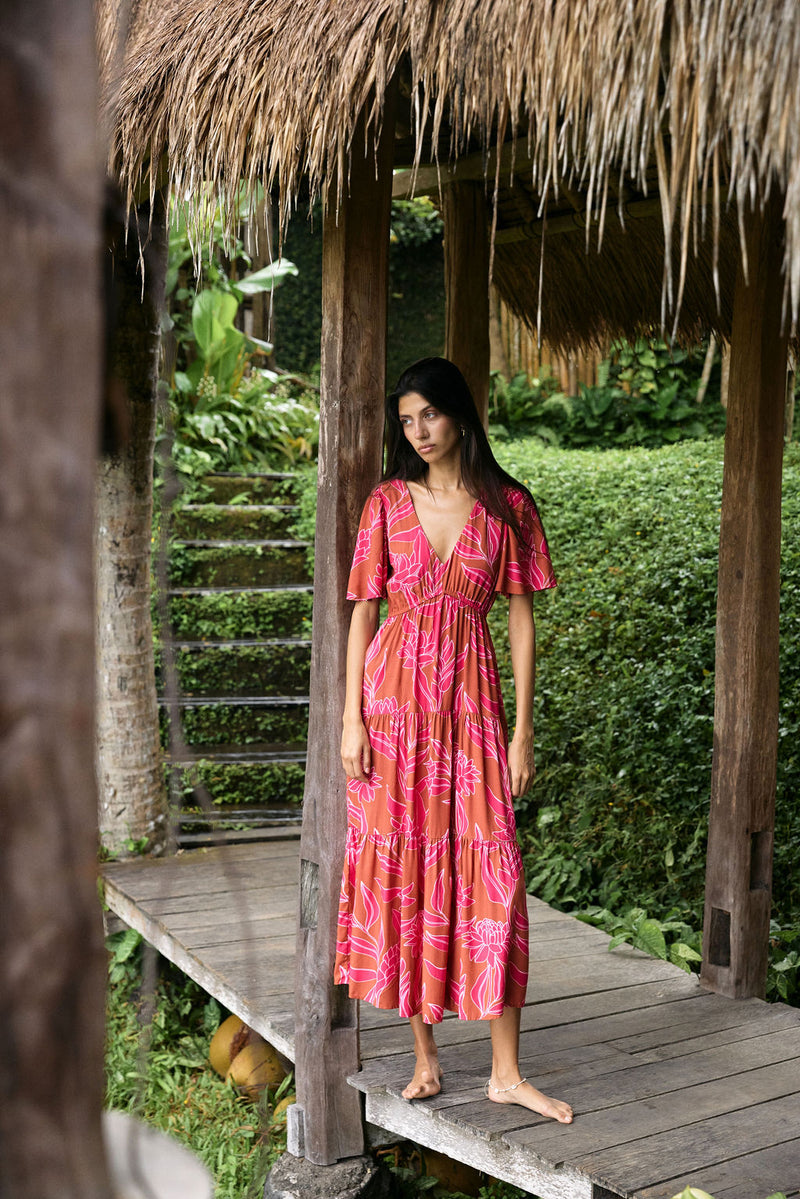 Woman with dark hair wearing tiered maxi dress with sleeves. It is the Mariana Dress in the Garden 'Awapuhi print. 