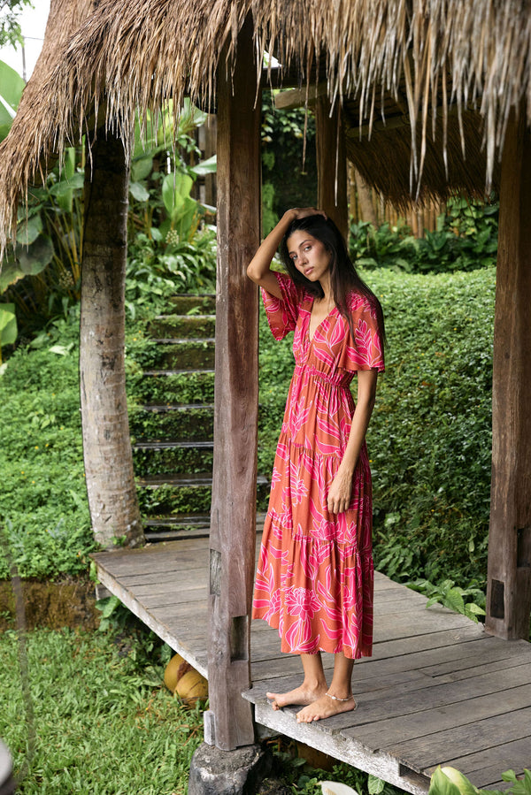 Woman with dark hair wearing tiered maxi dress with sleeves. It is the Mariana Dress in the Garden 'Awapuhi print. 