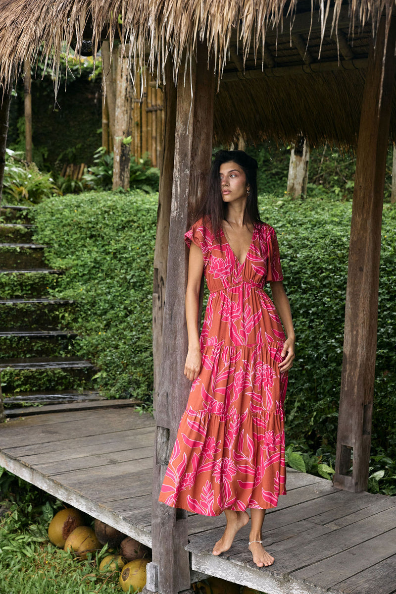 Woman with dark hair wearing tiered maxi dress with sleeves. It is the Mariana Dress in the Garden 'Awapuhi print. 