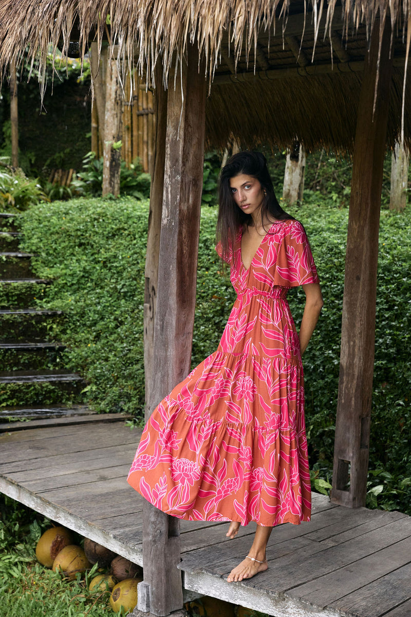 Woman with dark hair wearing tiered maxi dress with sleeves. It is the Mariana Dress in the Garden 'Awapuhi print. 