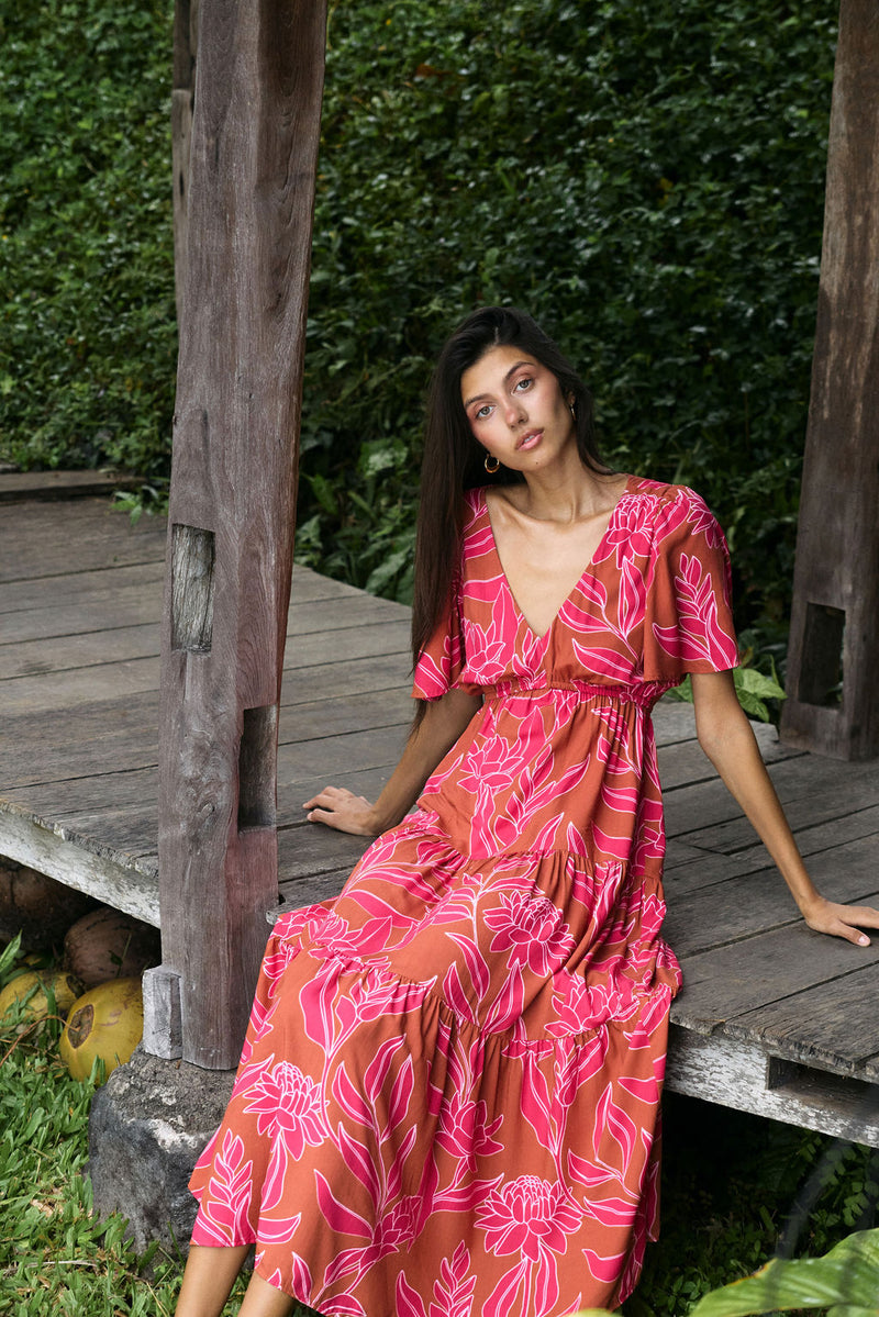 Woman with dark hair wearing tiered maxi dress with sleeves. It is the Mariana Dress in the Garden 'Awapuhi print. 