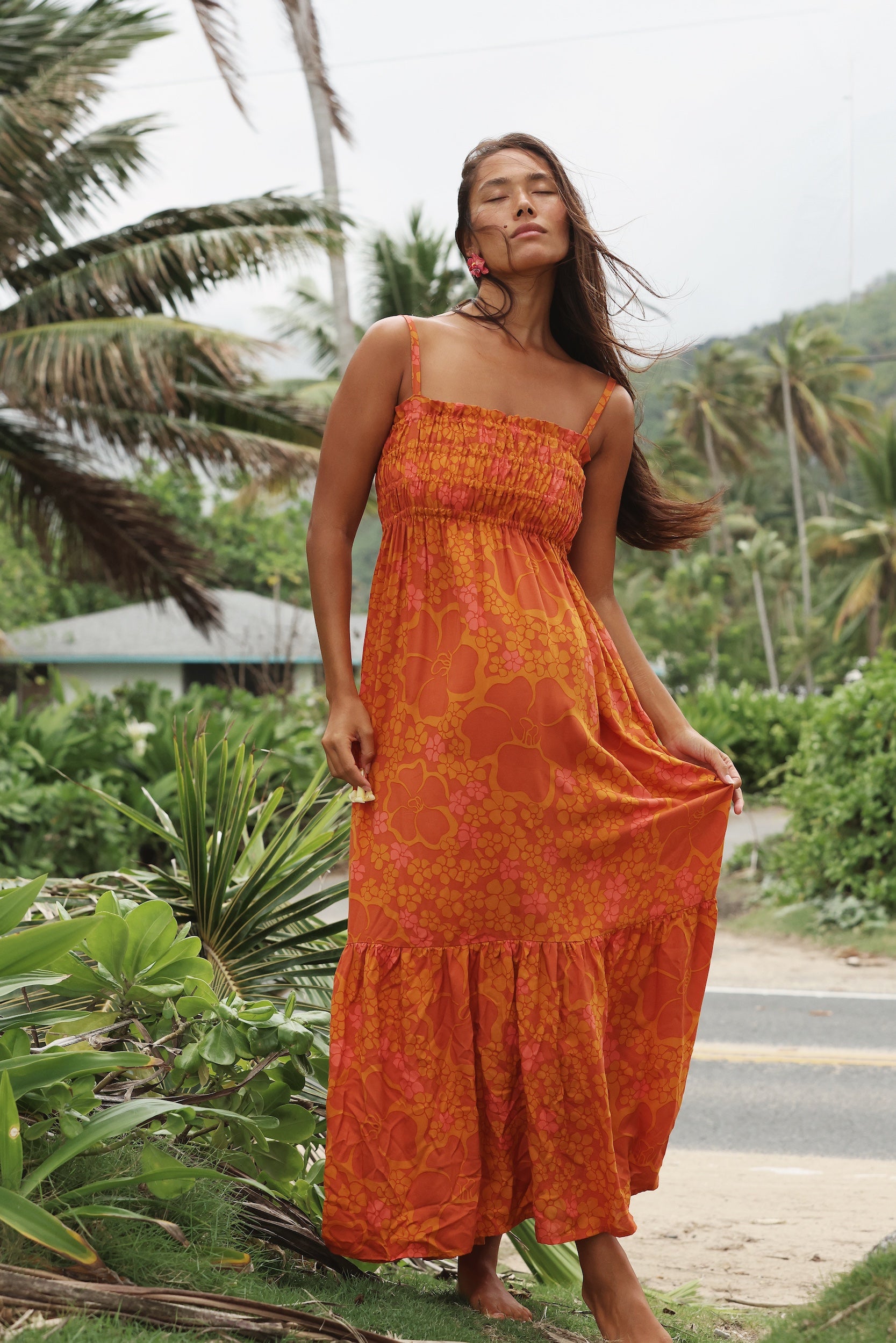 woman with brown hair wearing an orange long maxi dress with smocked bodice and spaghetti straps with puakenikeni print