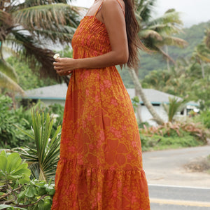 woman with brown hair wearing an orange long maxi dress with smocked bodice and spaghetti straps with puakenikeni print