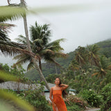 woman with brown hair wearing an orange long maxi dress with smocked bodice and spaghetti straps with puakenikeni print