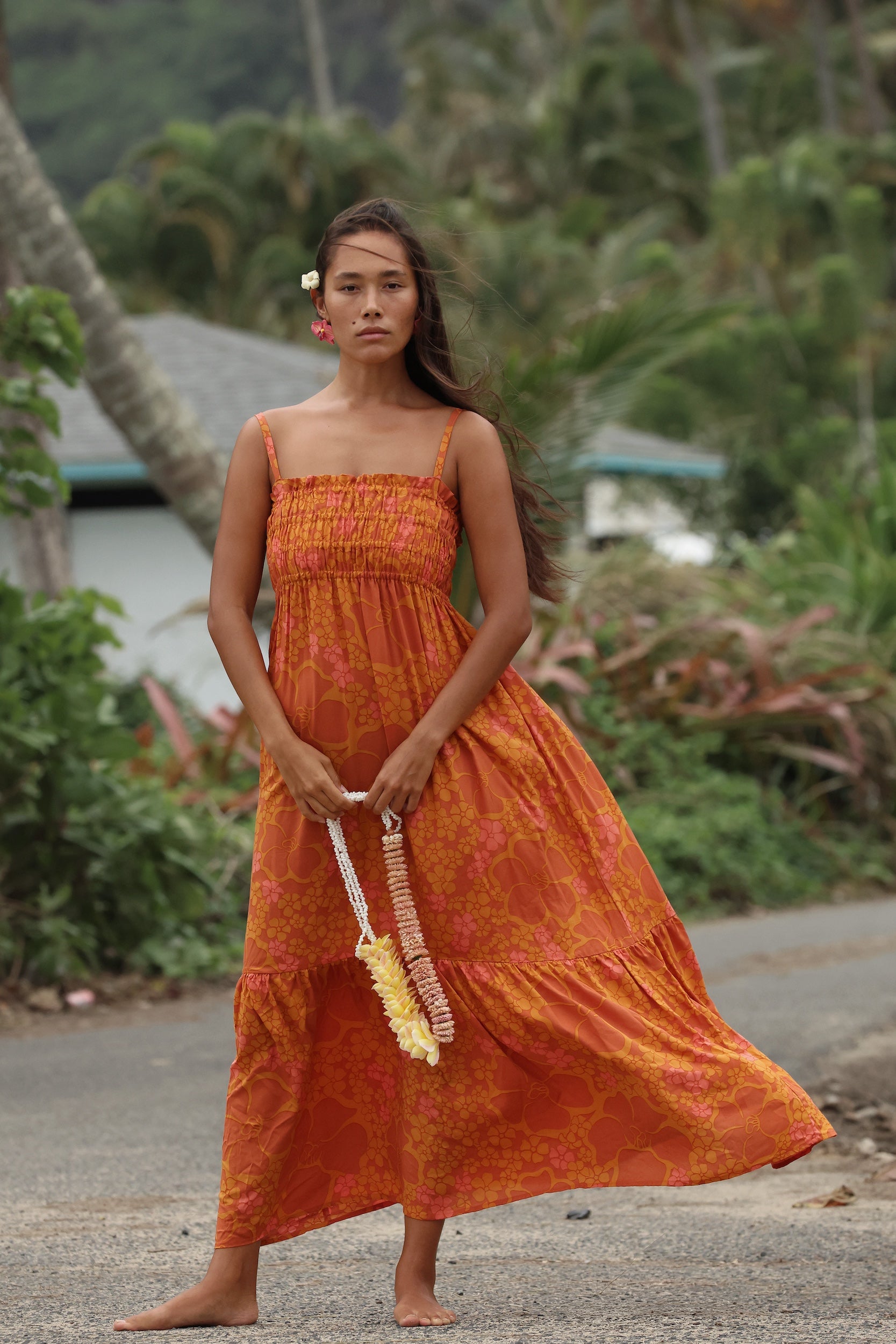 woman with brown hair wearing an orange long maxi dress with smocked bodice and spaghetti straps with puakenikeni print